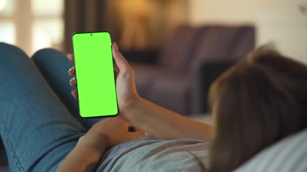 Γυναίκα στο σπίτι ξαπλωμένη σε καναπέ και χρησιμοποιώντας smartphone με πράσινη οθόνη mock-up σε κάθετη λειτουργία. Κορίτσι περιήγηση στο Internet, παρακολουθώντας περιεχόμενο, βίντεο, blogs. - Πλάνα, βίντεο