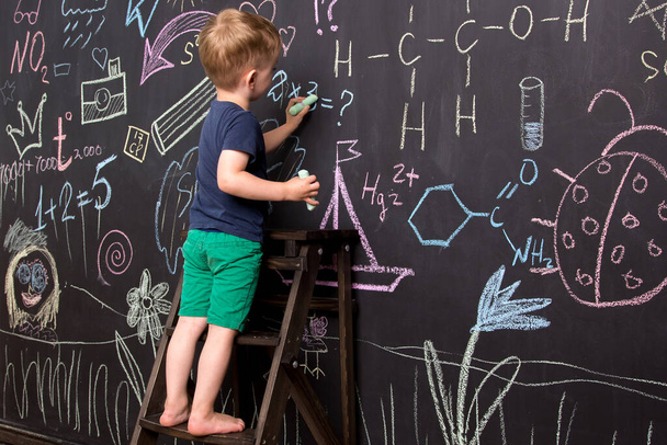 Kleine jongen tekent met kleurrijk krijt op een groot schoolbord. zwarte muur met kindertekeningen en formules.  - Foto, afbeelding