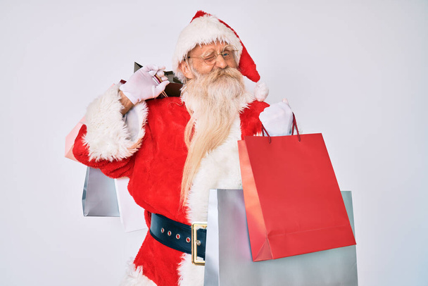 Staruszek z siwymi włosami i długą brodą w kostiumie Świętego Mikołaja trzymający mrugające torby na zakupy patrzące w kamerę z seksowną ekspresją, wesołą i szczęśliwą twarzą.  - Zdjęcie, obraz