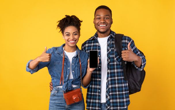 スマートフォンの画面を示すアフリカの旅行者のカップル親指アップ、黄色の背景をジェスチャー - 写真・画像
