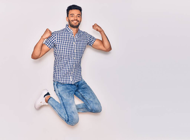 Young handsome hispanic man wearing casual clothes smiling happy. Jumping with smile on face celebrating with fists up over isolated white background - Foto, Imagem