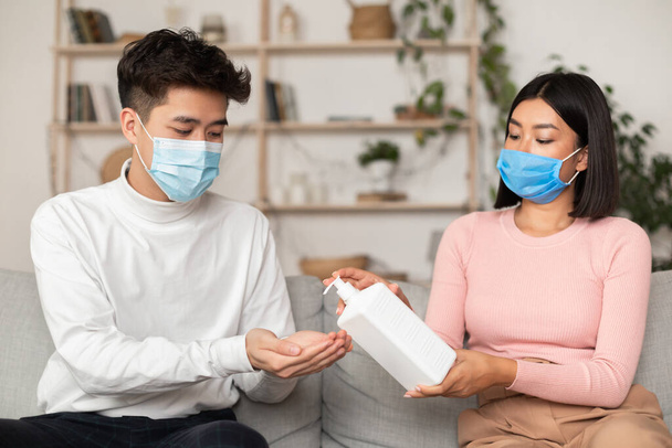 Chinese Couple Wearing Medical Masks Using Sanitizer Sitting At Home - Foto, Imagen