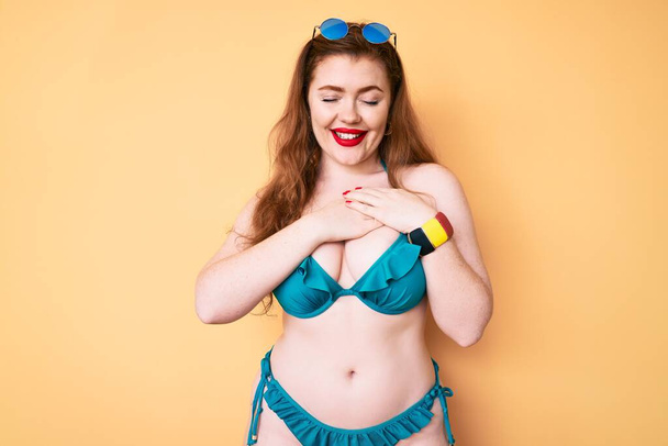 Young redhead woman wearing bikini smiling with hands on chest with closed eyes and grateful gesture on face. health concept.  - Photo, Image