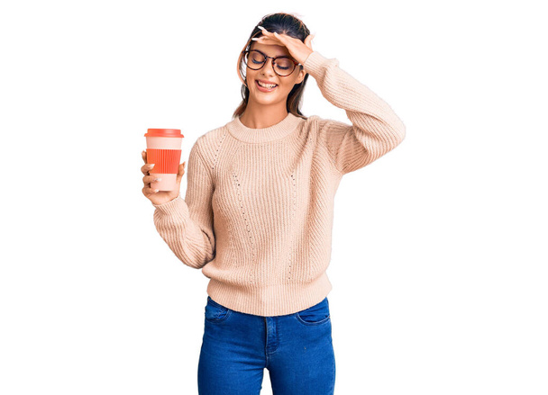 Jeune belle femme portant des lunettes tenant à emporter tasse de café stressé et frustré avec la main sur la tête, visage surpris et en colère  - Photo, image