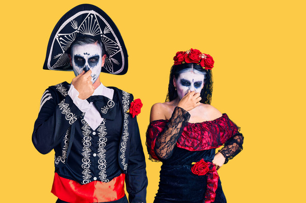 Young couple wearing mexican day of the dead costume over background smelling something stinky and disgusting, intolerable smell, holding breath with fingers on nose. bad smell  - Fotografie, Obrázek
