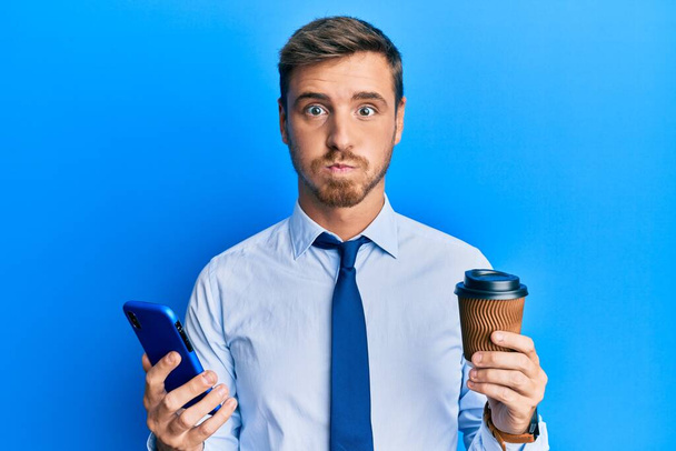 Όμορφος καυκάσιος επιχειρηματίας που χρησιμοποιεί smartphone και πίνει ένα φλιτζάνι καφέ φουσκώνοντας μάγουλα με αστείο πρόσωπο. στόμα φουσκωμένο με αέρα, που πιάνει αέρα.  - Φωτογραφία, εικόνα