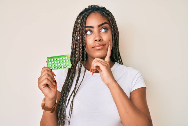 Mladá africká Američanka s copánky, které drží antikoncepci, vážná tvář přemýšlející o otázce s rukou na bradě, zamyšlená nad matoucím nápadem  - Fotografie, Obrázek