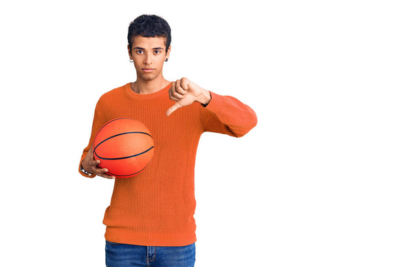 Young african amercian man holding basketball ball with angry face, negative sign showing dislike with thumbs down, rejection concept  - Φωτογραφία, εικόνα