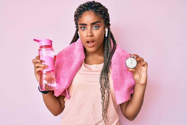 Молода афроамериканська спортивна жінка з колючками, що тримають пляшку з водою і годинник у шоці, виглядає скептично і саркастично, здивована відкритим ротом.  - Фото, зображення