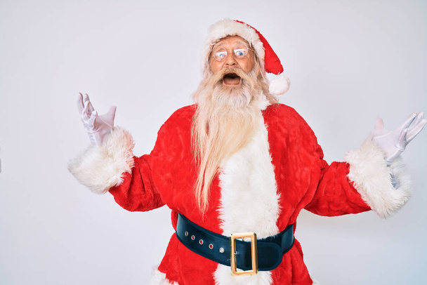 Old senior man with grey hair and long beard wearing santa claus costume celebrating mad and crazy for success with arms raised and closed eyes screaming excited. winner concept  - 写真・画像