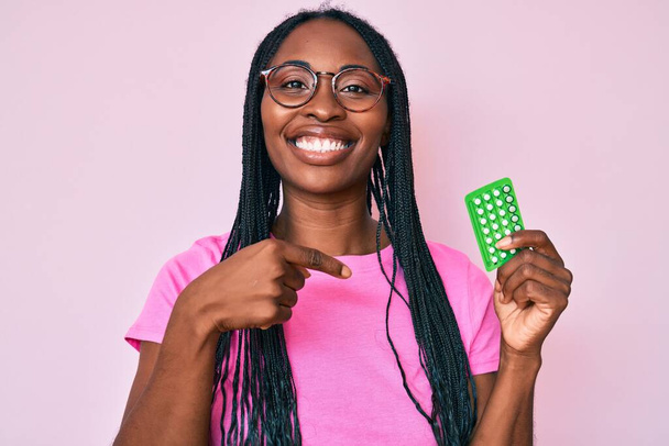 Африканська американка з колючками, що тримають протизаплідні таблетки, вказує пальцем на усмішку щасливою і гордою.  - Фото, зображення