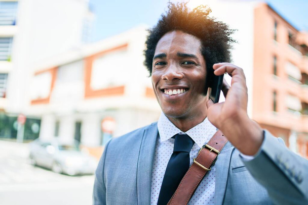 Mladý africký americký obchodník v obleku se šťastně usmívá. Stojíte s úsměvem na tváři a bavíte se na smartphonu na ulici. - Fotografie, Obrázek