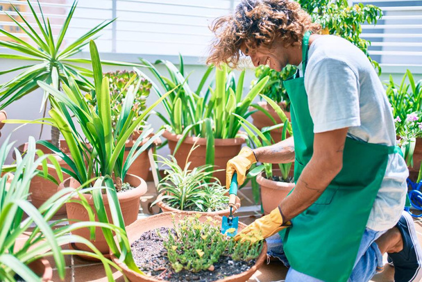 Молодой латиноамериканец улыбается счастливым ухоженным растениям с помощью садовой лопаты на террасе - Фото, изображение
