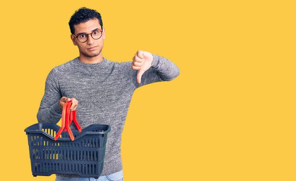 Spanyol jóképű fiatalember kezében szupermarket bevásárlókosár dühös arc, negatív jel mutatja, nem tetszik hüvelykujjal lefelé, elutasítás koncepció  - Fotó, kép