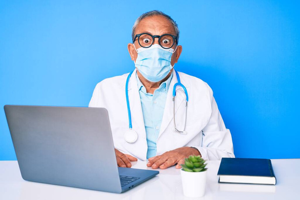 Starszy przystojny mężczyzna z siwymi włosami noszący mundur lekarza i maskę medyczną przestraszony i zdumiony otwartymi ustami dla zaskoczenia, niedowierzanie twarz  - Zdjęcie, obraz