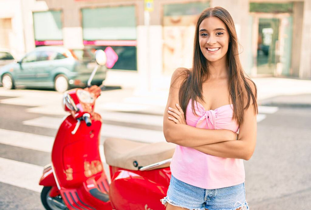 Νεαρή Ισπανίδα γυναίκα χαμογελά ευτυχισμένη στέκεται πάνω από σκούτερ στην πόλη - Φωτογραφία, εικόνα