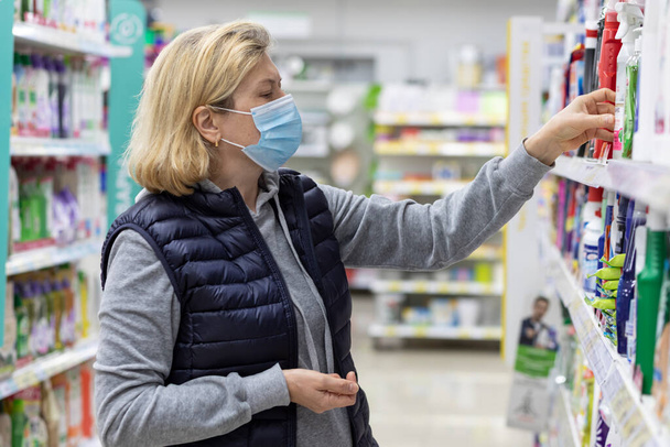 Una anciana con una máscara facial hace una compra en el departamento doméstico de un supermercado. - Foto, imagen
