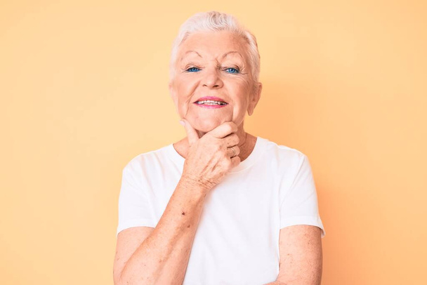 Eine ältere schöne Frau mit blauen Augen und grauen Haaren trägt ein klassisches weißes T-Shirt vor gelbem Hintergrund und blickt selbstbewusst in die Kamera, die Arme verschränkt und die Hand am Kinn erhoben. Positives Denken.  - Foto, Bild