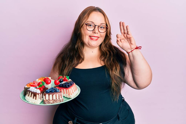 Además tamaño caucásico joven mujer sosteniendo rebanadas de pastel haciendo signo de ok con los dedos, sonriendo gesto amistoso excelente símbolo  - Foto, imagen