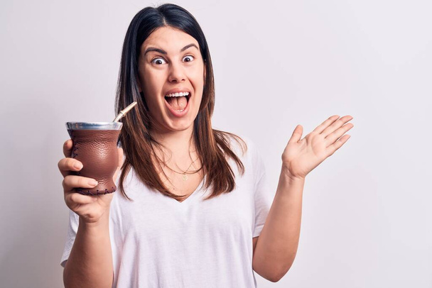 Young beautiful brunette woman drinking mate infusion beverage over white background celebrating achievement with happy smile and winner expression with raised hand - Foto, imagen