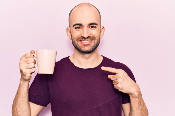 Young handsome bald man drinking cup of coffee smiling happy pointing with hand and finger  - Foto, imagen