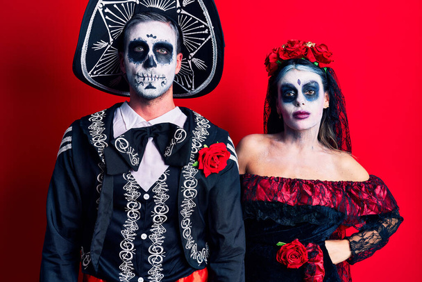Casal jovem vestindo dia mexicano do traje morto sobre vermelho relaxado com expressão séria no rosto. simples e natural olhando para a câmera.  - Foto, Imagem