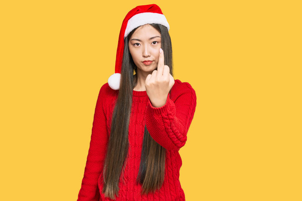 Young chinese woman wearing christmas hat showing middle finger, impolite and rude fuck off expression  - Photo, image