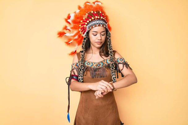 Young beautiful latin girl wearing indian costume checking the time on wrist watch, relaxed and confident  - 写真・画像