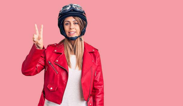 Młoda piękna kobieta w kasku motocyklowym uśmiechnięta ze szczęśliwą twarzą mrugająca do kamery robiąc znak zwycięstwa. numer dwa..  - Zdjęcie, obraz
