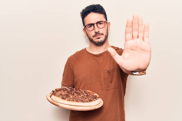 Jeune homme hispanique portant des lunettes tenant une pizza italienne à la main ouverte faisant signe d'arrêt avec expression sérieuse et confiante, geste de défense  - Photo, image