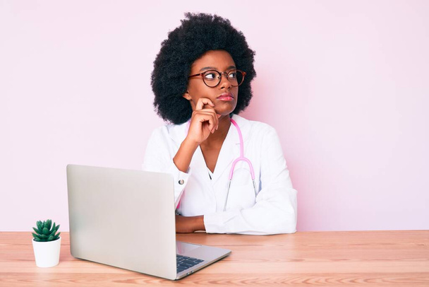 Mujer afroamericana joven que usa estetoscopio médico trabajando con computadora portátil cara seria pensando en la pregunta con la mano en la barbilla, reflexivo acerca de la idea confusa  - Foto, imagen