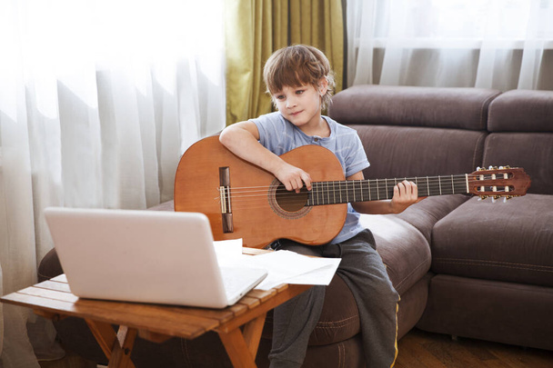 Kid boy playing guitar and watching online lessons on laptop while practicing at home. Stay home. quarantine. Online training, online classes. - Фото, изображение