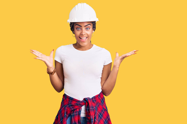 Jeune femme afro-américaine avec des tresses portant un casque et des vêtements de constructeur célébrant la victoire avec un sourire heureux et l'expression gagnante avec les mains levées  - Photo, image