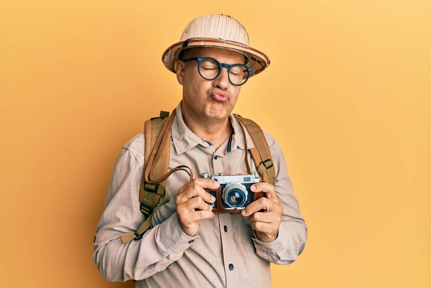Łysy mężczyzna w średnim wieku, noszący kapelusz odkrywcy i zabytkową kamerę, patrzący w kamerę i dmuchający pocałunek, jest uroczy i seksowny. wyrażenie miłości.  - Zdjęcie, obraz