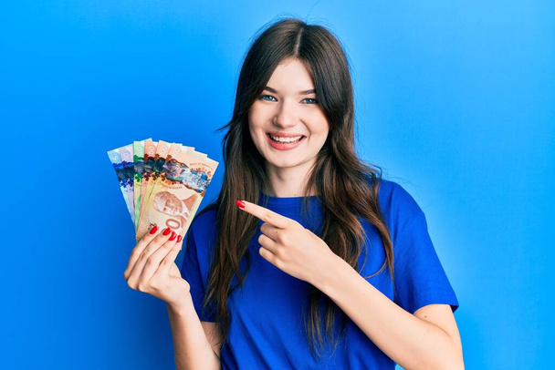 Young beautiful caucasian girl holding canadian dollars smiling happy pointing with hand and finger  - Fotoğraf, Görsel