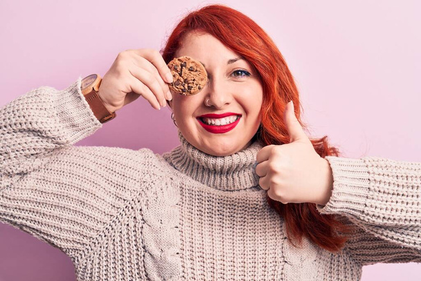 Mladý krásný ryšavý žena stojící drží čokoládové sušenky přes oko usměvavý šťastný a pozitivní, palec nahoru dělá vynikající a schvalovací znamení - Fotografie, Obrázek