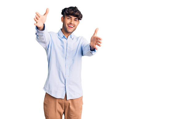 Junger hispanischer Mann in lässigem Hemd, der mit offenen Armen in die Kamera lächelt und sich umarmt. fröhlicher Ausdruck, der das Glück umarmt.  - Foto, Bild