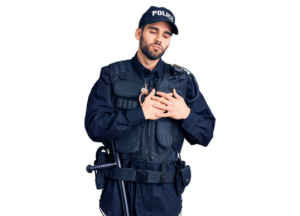 Junger gutaussehender Mann mit Bart in Polizeiuniform, lächelnd, die Hände auf der Brust, mit geschlossenen Augen und dankbarer Geste im Gesicht. Gesundheitskonzept.  - Foto, Bild