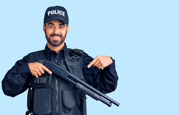 Joven hispano vistiendo uniforme policial sosteniendo escopeta sonriendo feliz señalando con la mano y el dedo  - Foto, Imagen