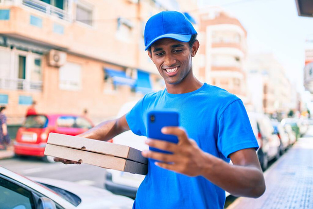Afrikaanse bezorger draagt koerier uniform buiten met behulp van smartphone en het houden van meeneem pizza dozen - Foto, afbeelding
