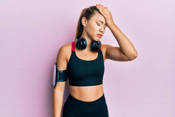 Beautiful young blonde woman wearing gym clothes and using headphones surprised with hand on head for mistake, remember error. forgot, bad memory concept.  - Foto, imagen