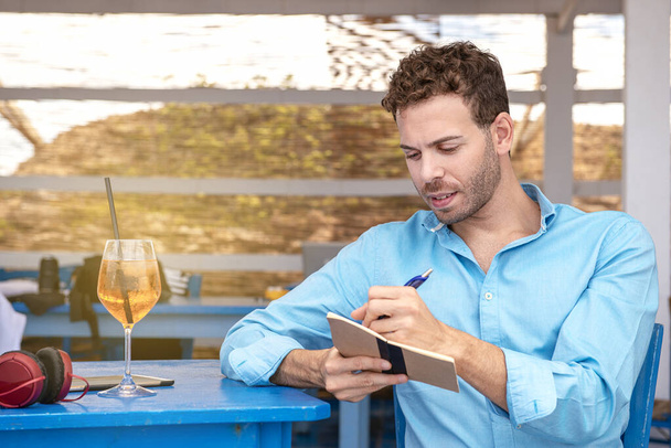 Levoruký mladý muž sedící v baru na pláži a psal svůj domácí úkol o chytrou práci venku - Fotografie, Obrázek