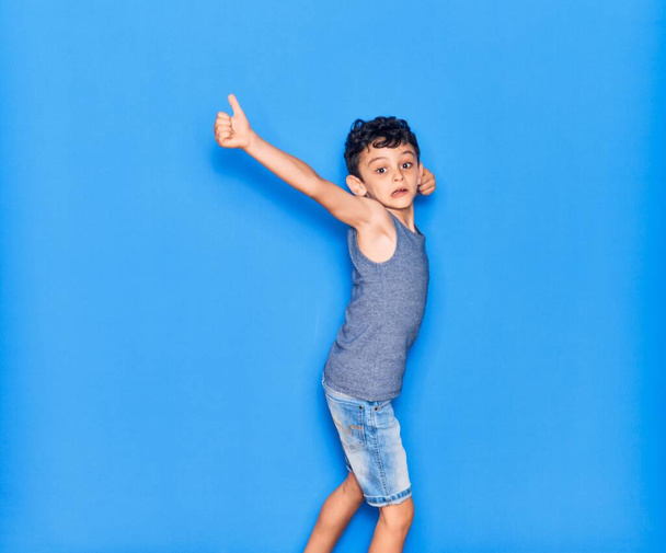 Αξιολάτρευτο παιδί που φοράει καθημερινά ρούχα. Άλμα κάνει εντάξει σημάδι με τον αντίχειρα πάνω από απομονωμένο μπλε φόντο - Φωτογραφία, εικόνα