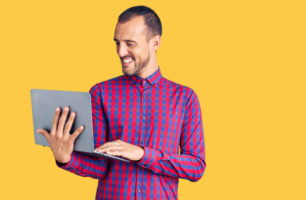 Молодой красивый мужчина, работающий с компьютером ноутбук выглядит позитивным и счастливым стоя и улыбаясь с уверенной улыбкой показывая зубы  - Фото, изображение