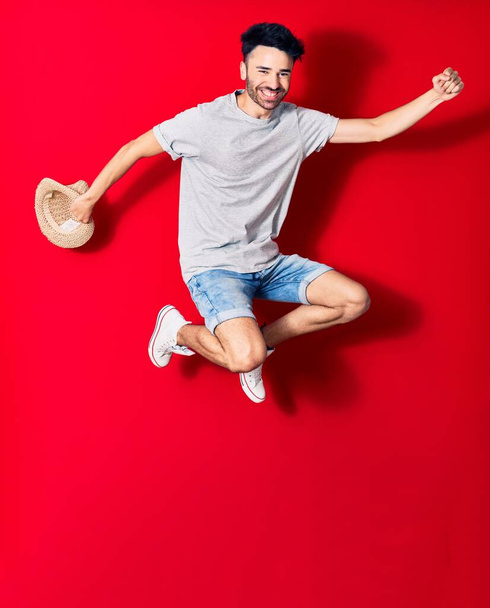 Νεαρός όμορφος Ισπανός που φοράει καθημερινά ρούχα χαμογελώντας χαρούμενος. Άλμα με χαμόγελο στο πρόσωπο κρατώντας το καλοκαιρινό καπέλο γιορτάζει με γροθιά πάνω από απομονωμένο κόκκινο φόντο. - Φωτογραφία, εικόνα