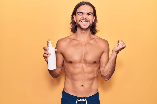 Young handsome man wearing swimwear holding sunscreen lotion screaming proud, celebrating victory and success very excited with raised arm  - Photo, Image