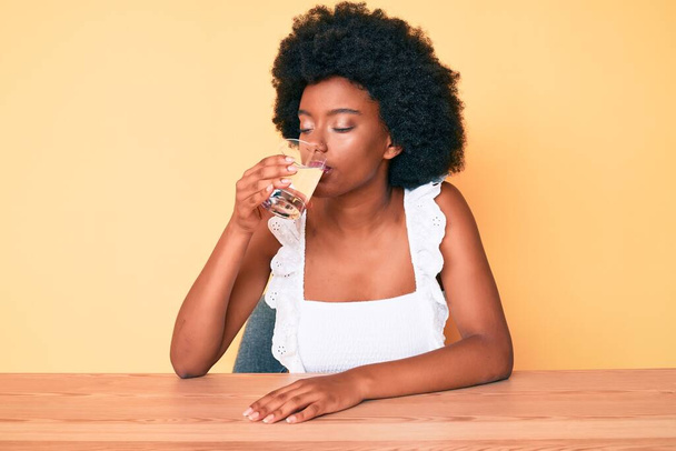 Afro saçlı genç Afrikalı Amerikalı kadın evdeki masada oturmuş bir bardak tatlı su içiyor. - Fotoğraf, Görsel