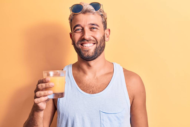 Giovane uomo biondo bello con la barba in vacanza bere bicchiere di succo d'arancia sano guardando positivo e felice in piedi e sorridente con un sorriso fiducioso che mostra i denti - Foto, immagini