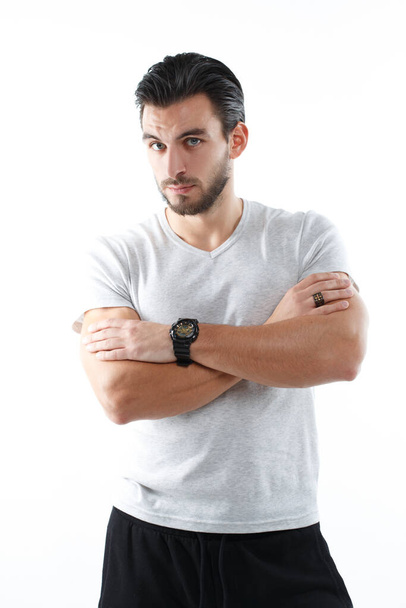 A charismatic young athletic man in a light tight shirt stands with arms crossed over his chest isolated on a white background. - Foto, immagini