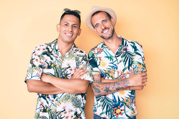 夏の帽子をかぶっている2人の男性とハワイのシャツを着た若いゲイのカップルは、カメラを見て両腕を組んで笑顔。正の人.  - 写真・画像
